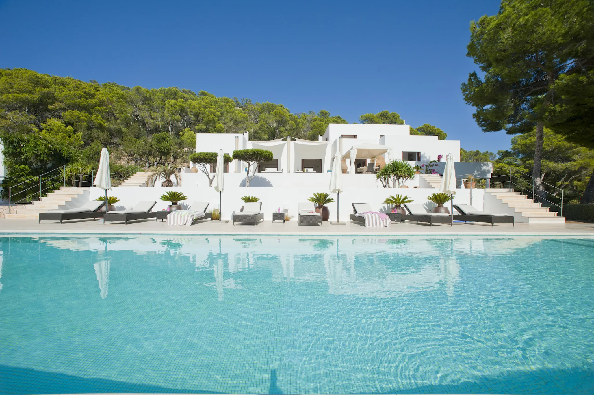 A large white villa featuring a swimming pool surrounded by lush greenery under a clear blue sky.
