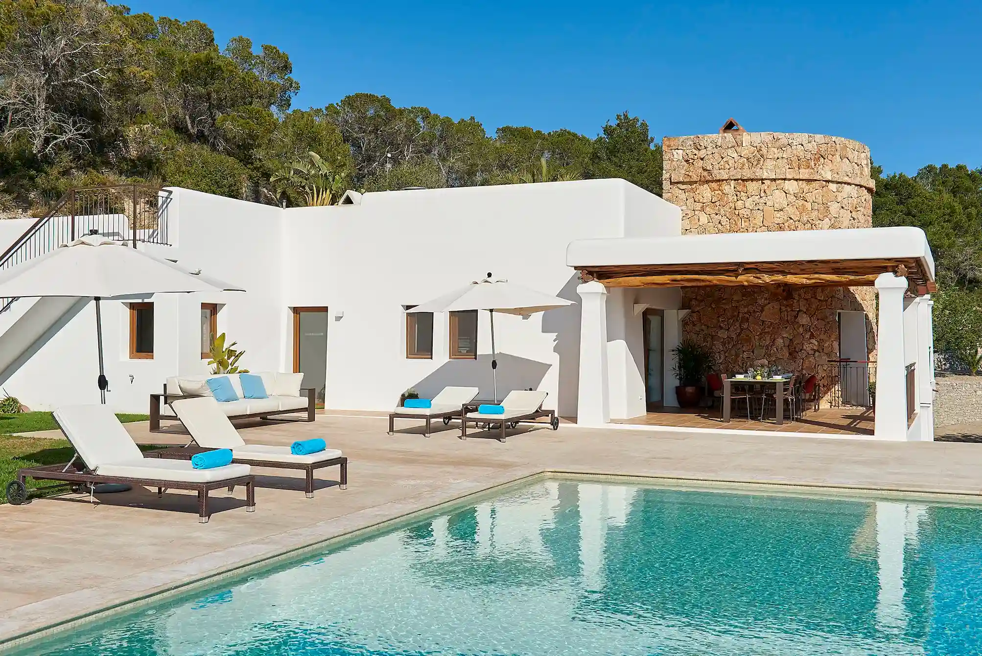 A villa featuring a swimming pool surrounded by sun loungers, set in a sunny outdoor environment.