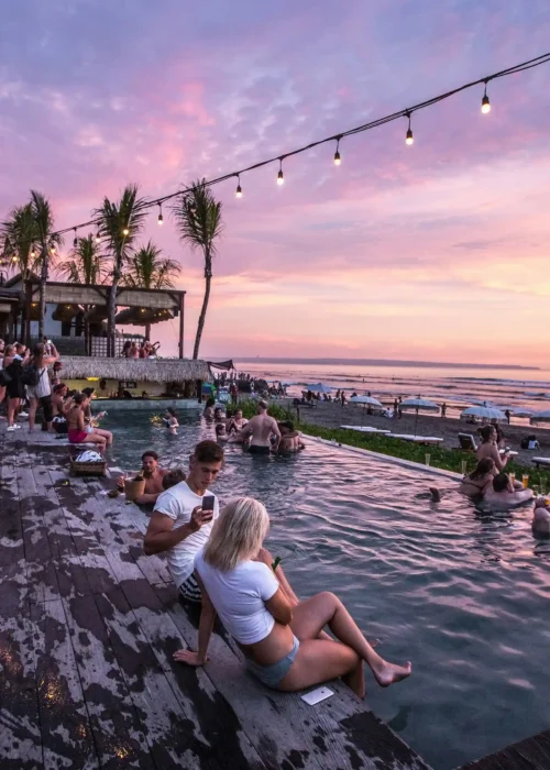 Individuals parting on the club next to the beach as the sun sets, casting warm hues across the water and surrounding area.